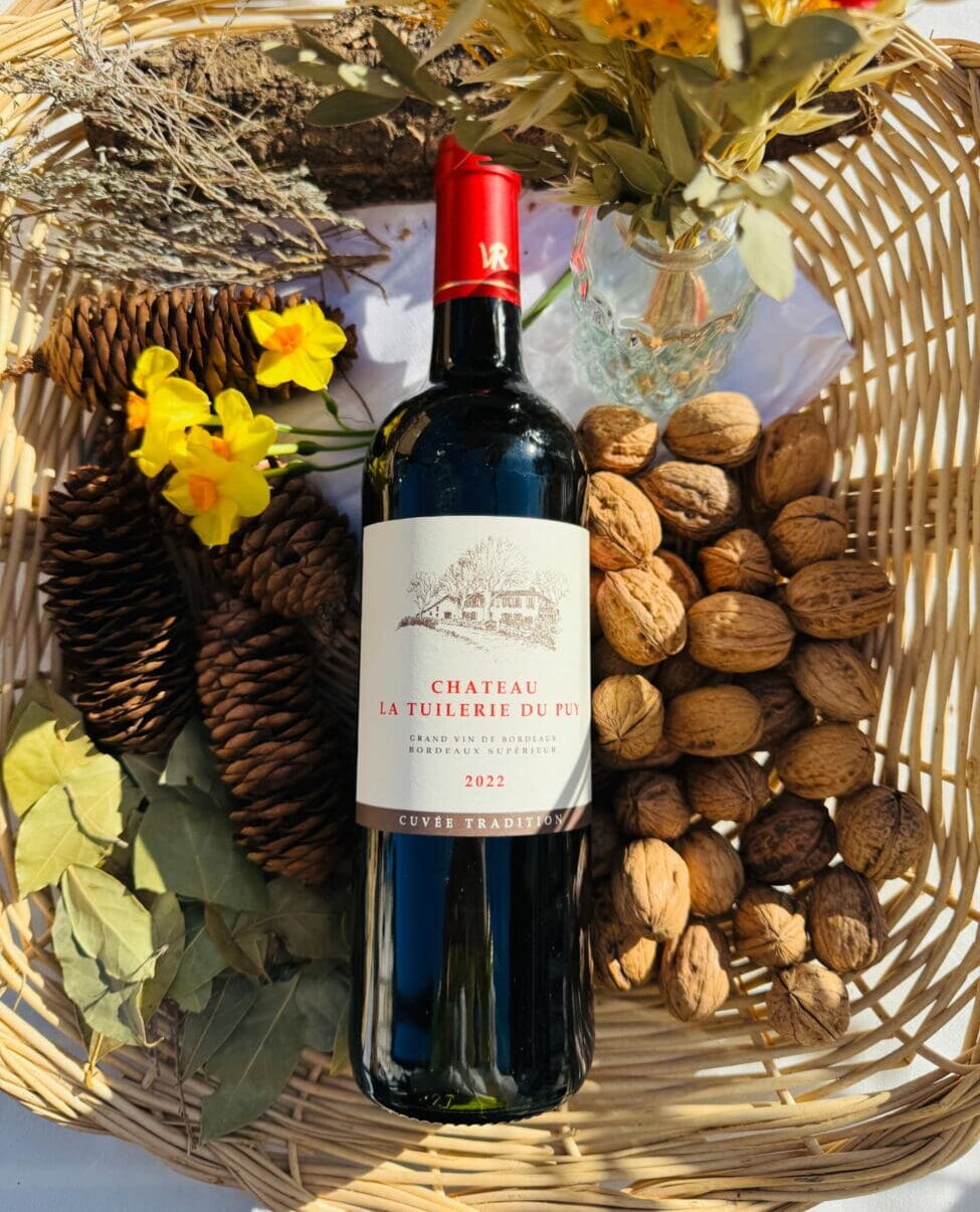 Bouteille de vin rouge Château La Tuilerie Du Puy "Cuvée Tradition" 2022 dans un panier en osier entourée de noix, de fleurs jaunes et de feuilles de laurier.