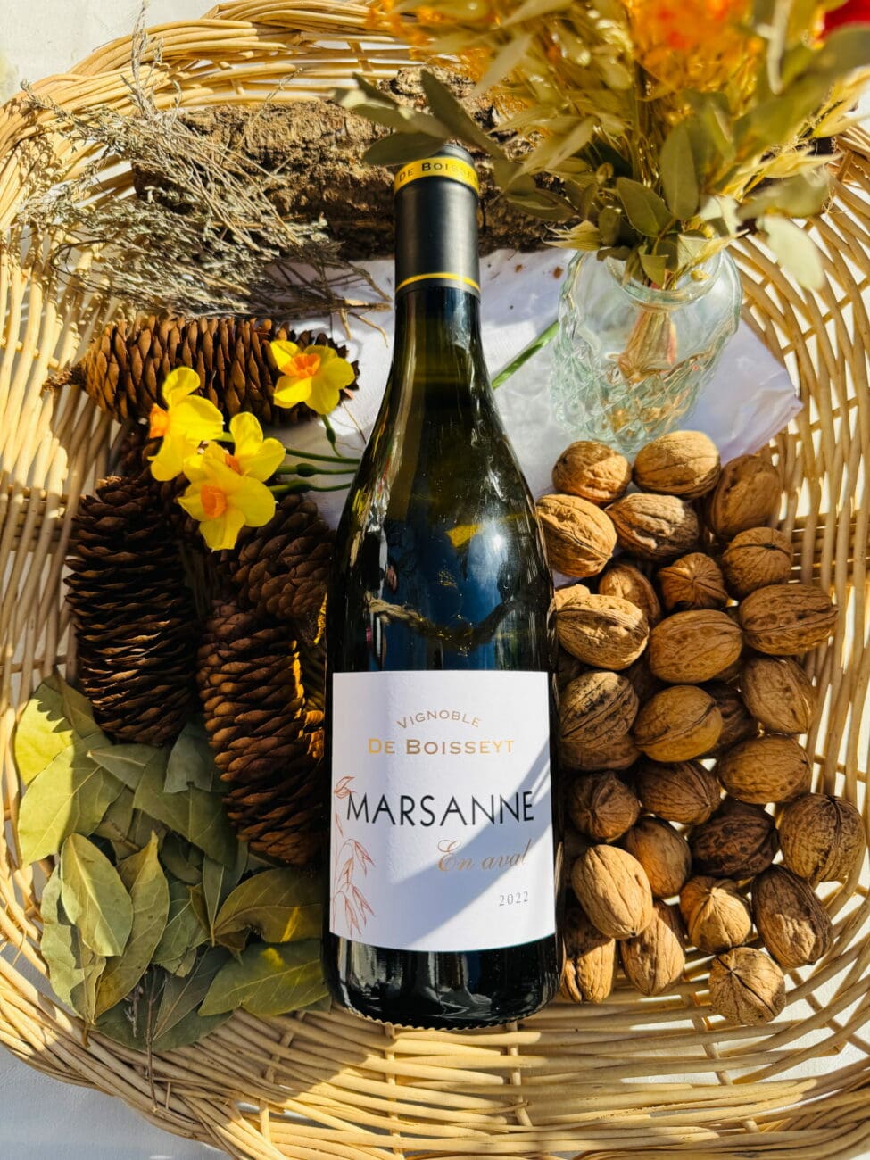 Bouteille de vin blanc Marsanne "En Aval" 2022 du Vignoble De Boisseyt dans un panier en osier entourée de noix, de pommes de pin et de fleurs jaunes.