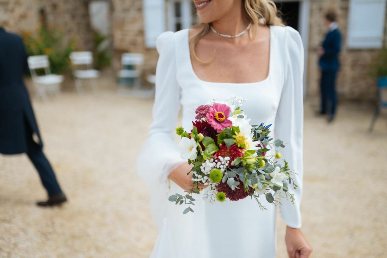 Un gros plan sur le buste d'une mariée portant une robe blanche à col carré et manches ballons, tenant un bouquet coloré de zinnia, cosmos et eucalyptus lors d'un mariage en extérieur à Dijon.