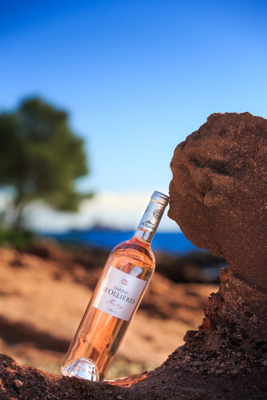 Bouteille de vin rosé Château d'Ollières Prestige appuyée contre un rocher rouge sous un ciel bleu azur.