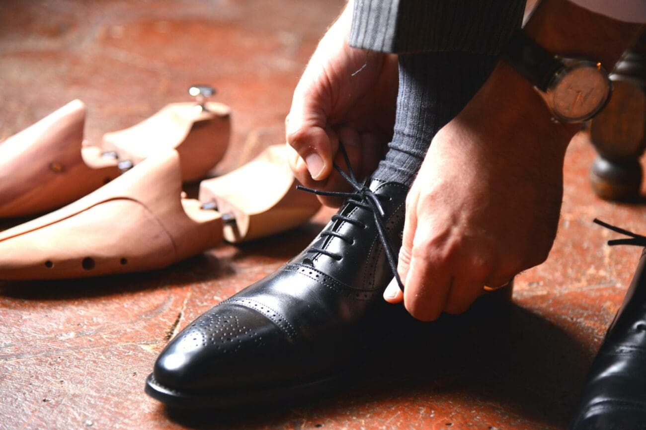 Gros plan sur un marié en chaussettes grises et pantalon de costume, laçant une de ses chaussures richelieu en cuir noir brillant sur un sol carrelé en terre cuite. Sa main porte une alliance dorée et une montre élégante est visible à son poignet.