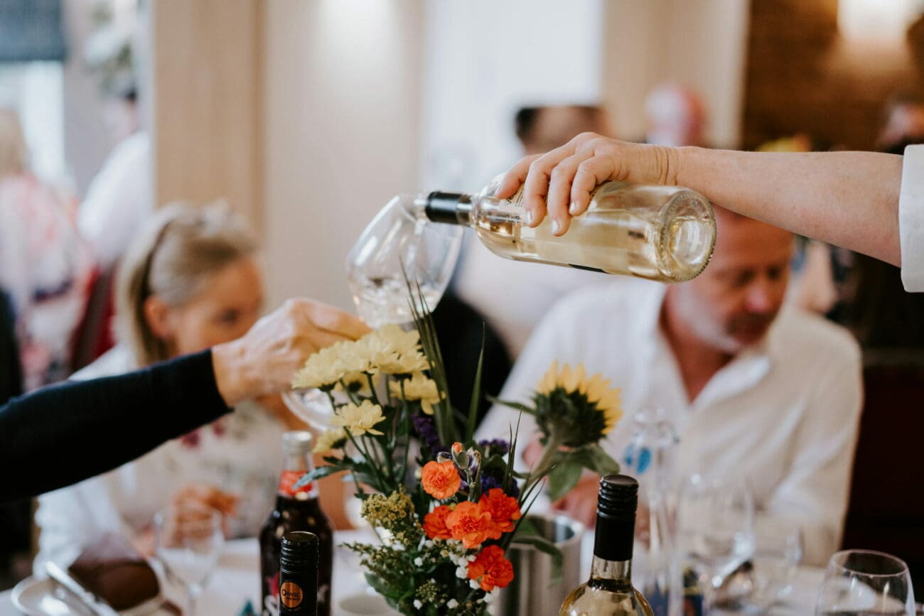 Un serveur verse du vin blanc d'une bouteille transparente dans un verre à pied lors d'un repas de mariage convivial.