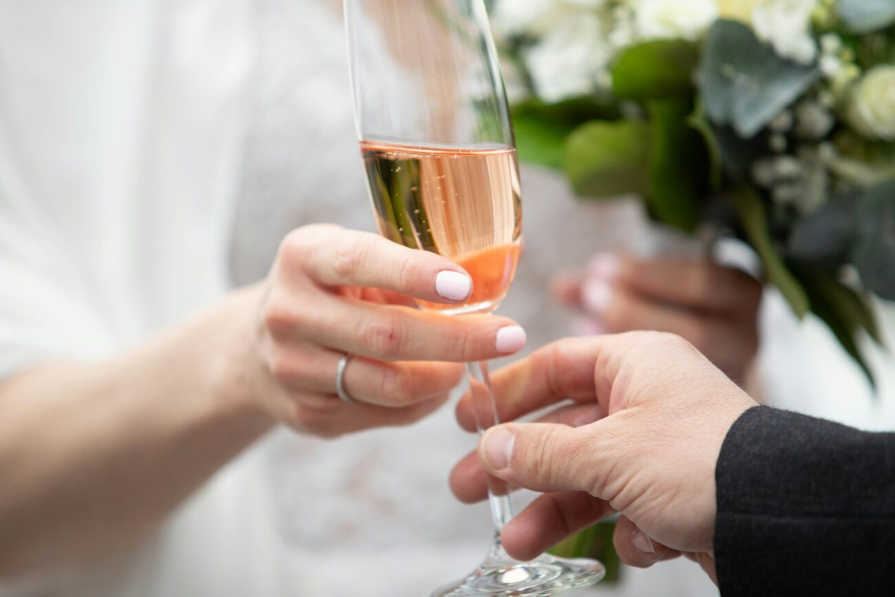 Gros plan sur une main d'homme s'apprêtant à prendre ou à trinquer avec une flûte de vin rosé tenue par une main de femme portant une alliance, lors d'un mariage.
