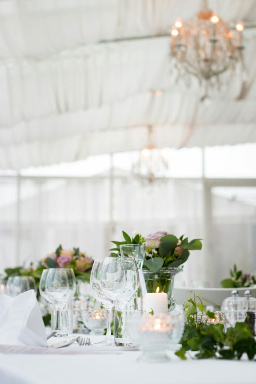 Gros plan sur une table de mariage sous un chapiteau blanc, décorée de verres à vin cristallins, de bougies allumées et de compositions florales champêtres.