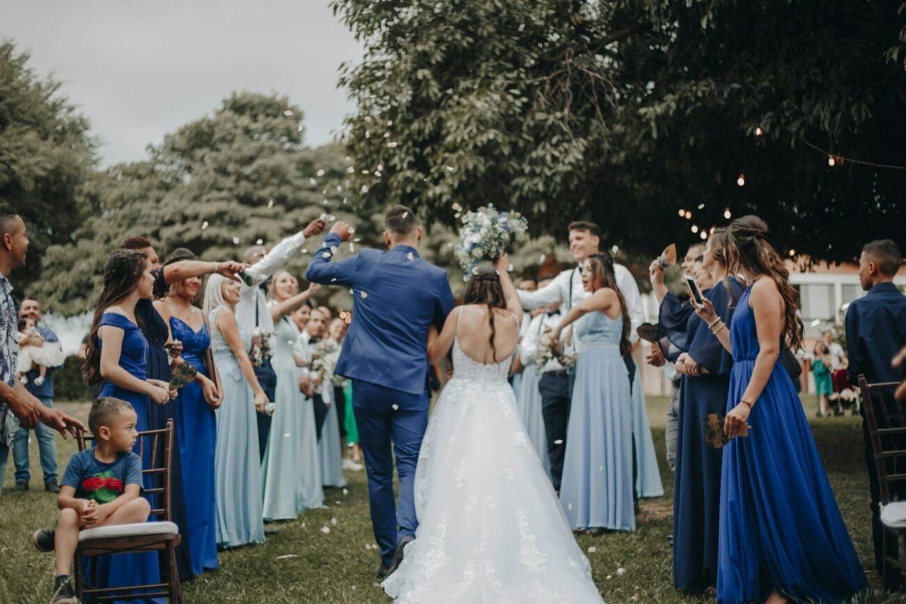 Haie d'honneur pour les mariés marchant vers leur réception. Demoiselles d'honneur en robes bleues et invités célébrant sous les arbres dans un jardin. Scène de mariage festive et conviviale.