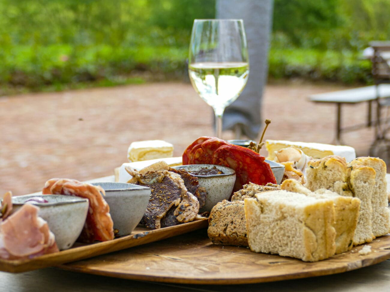 Le Happiness Manager de vins-mariages.net : un plateau dînatoire gourmand avec pain artisanal, charcuterie fine, fromages et un verre de vin blanc en arrière-plan lors d'une réception.