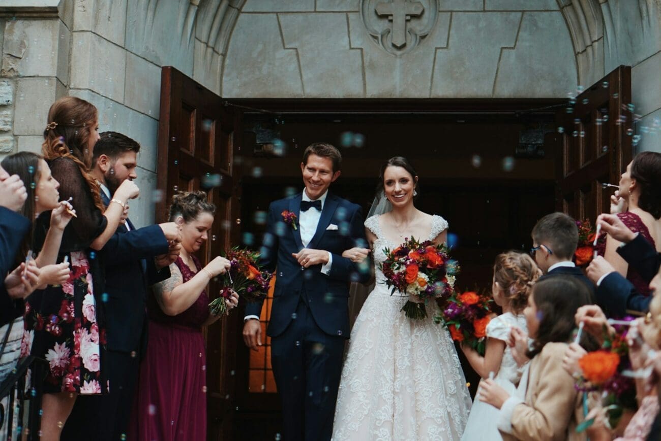 Sortie de cérémonie de mariage : les mariés heureux sont accueillis par des bulles de savon et leurs invités. La mariée porte une robe en dentelle et le marié un costume bleu marine. Scène festive avant le vin d'honneur.