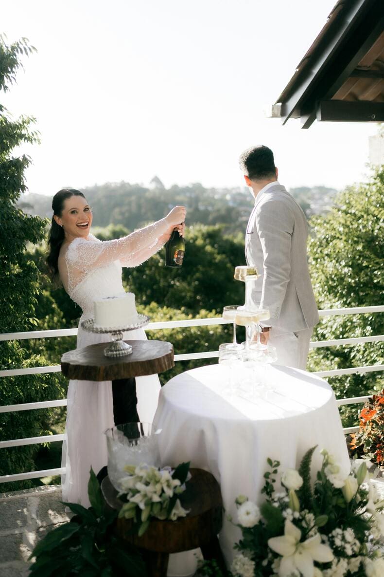 Mariée joyeuse ouvrant une bouteille de champagne à côté d'une pyramide de coupes sur une terrasse de mariage. Célébration festive en extérieur avec gâteau de mariage et décoration florale blanche.