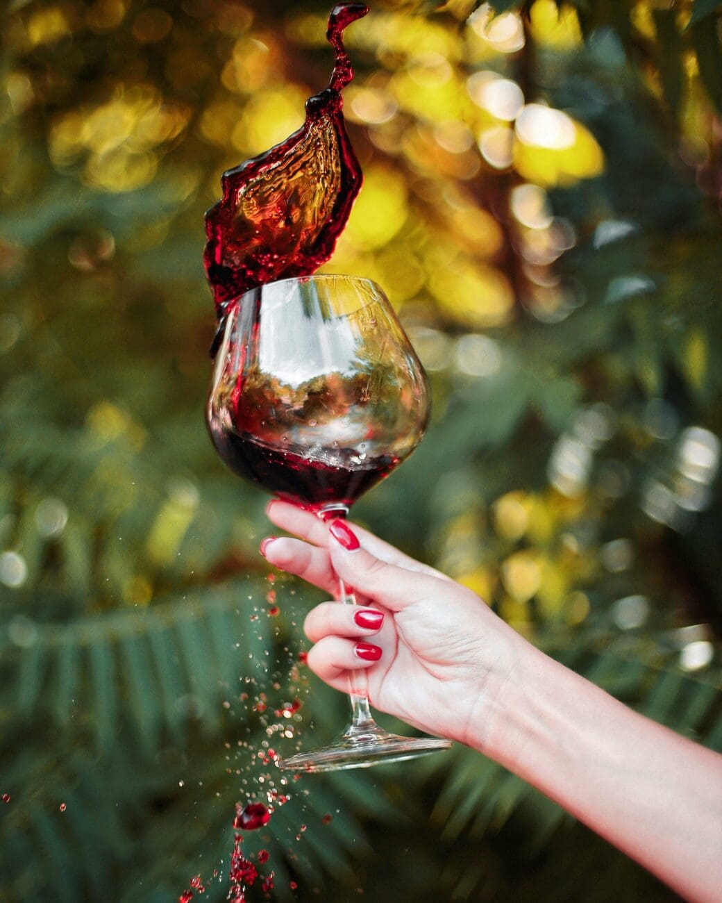 Gros plan sur une main de femme avec une manucure rouge vif, tenant fermement le pied d'un grand verre à vin ballon rempli de vin rouge. Du vin rouge jaillit énergiquement du verre, créant une vague de liquide rouge rubis. L'arrière-plan est un feuillage vert luxuriant très flou, évoquant une réception de mariage en plein air et ensoleillée. L'ambiance est vivante, spontanée et artistique.