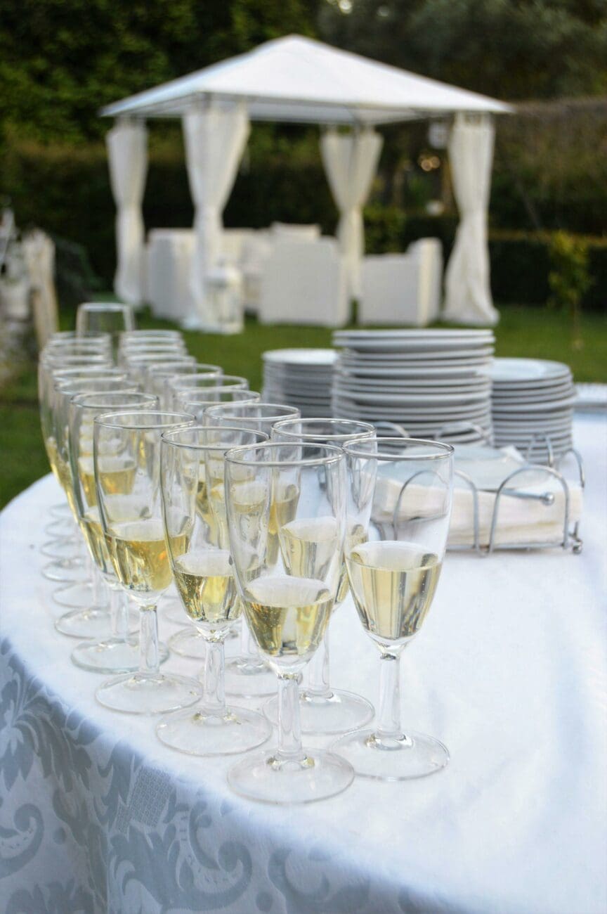 Gros plan de flûtes de champagne remplies et alignées sur une table de buffet avec nappe damassée, prête pour un vin d'honneur de mariage en extérieur. En arrière-plan, une tonnelle blanche élégante avec des voilages sur une pelouse. L'ambiance est festive et raffinée.