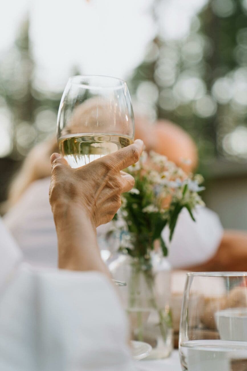 Gros plan sur une main tenant un verre de vin blanc à la robe dorée lors d'un déjeuner de mariage en extérieur. Décoration de table avec fleurs printanières en arrière-plan flou.