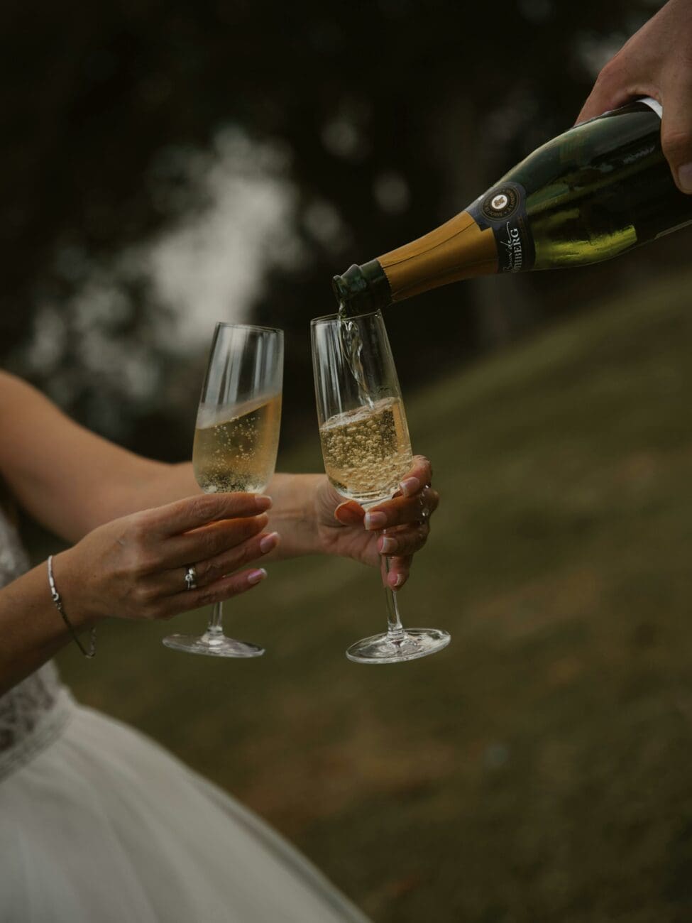 Service de bulles dans deux flûtes lors d'une réception de mariage en extérieur, focus sur la robe dorée et les bulles.