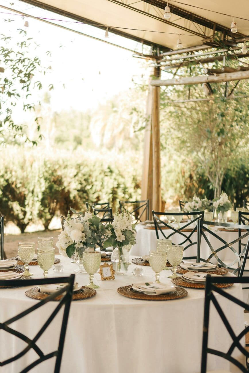 Vue d'une table de mariage rectangulaire sous une structure couverte avec guirlandes lumineuses de type guinguette. Décoration de table épurée avec chaises en fer forgé.