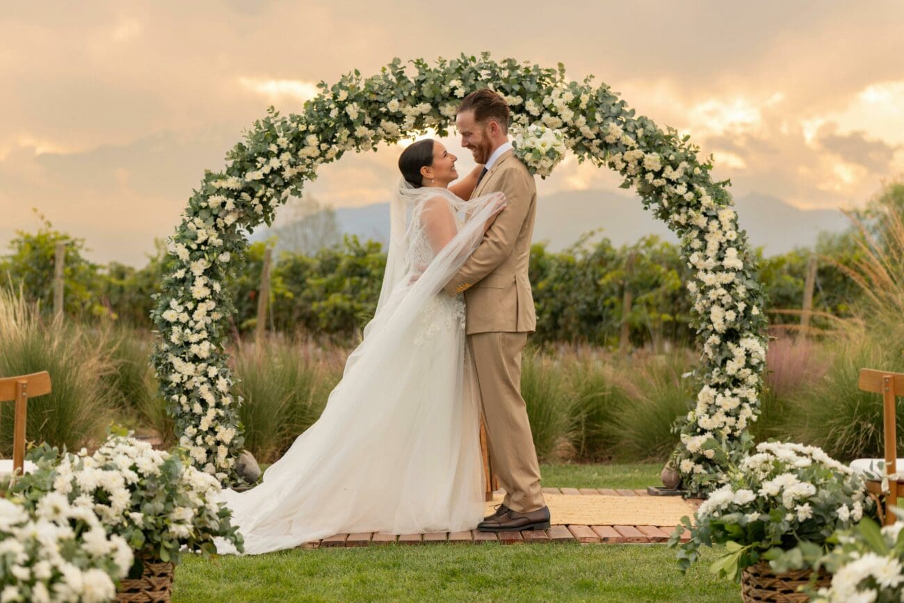 Couple de mariés s'enlaçant sous une arche florale circulaire dans un vignoble au coucher du soleil. Mariage en plein air dans les vignes avec vue sur les montagnes.