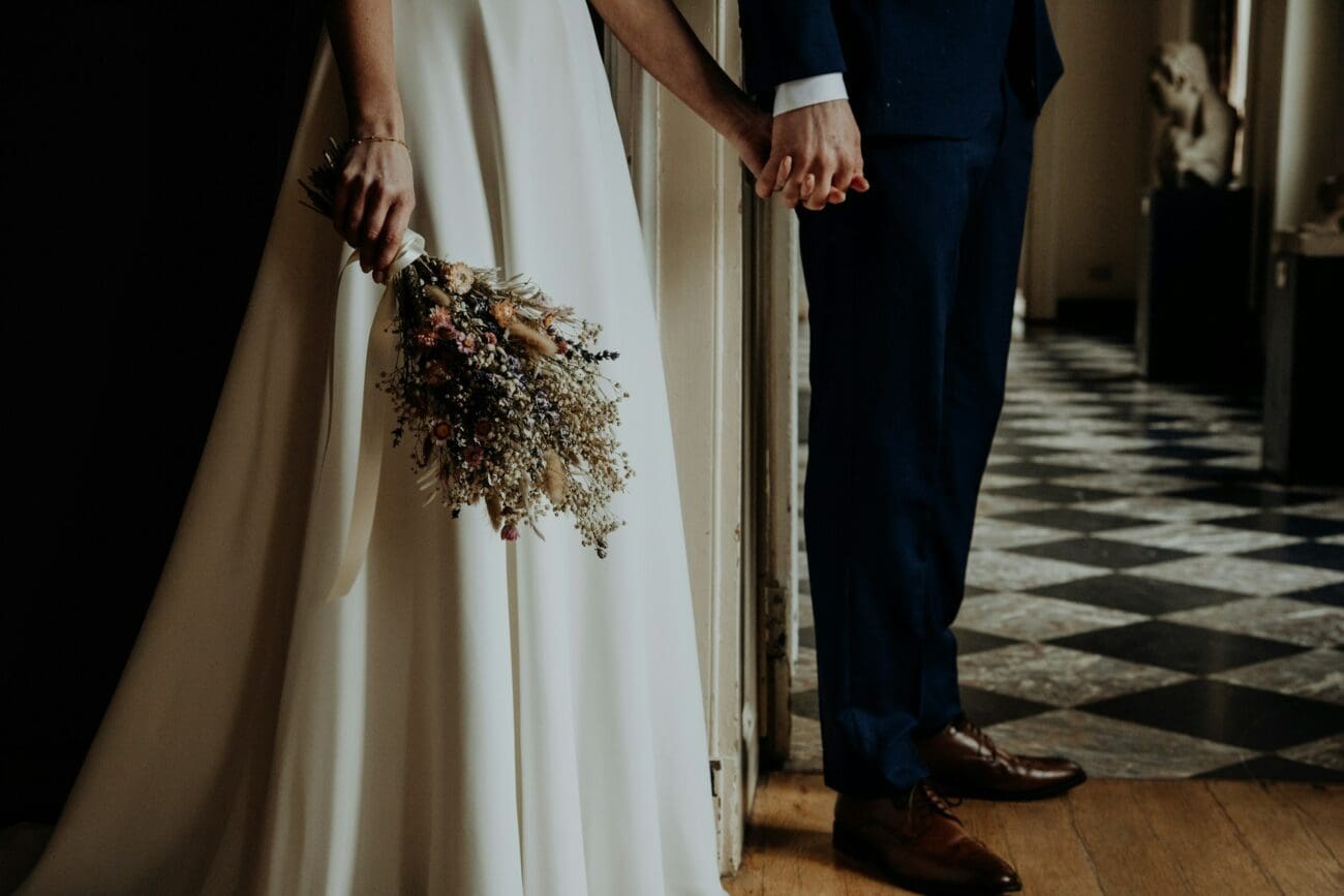 Vue recadrée d'un couple de mariés se tenant par la main, la mariée tenant un bouquet de fleurs séchées, sur un sol en damier classique.