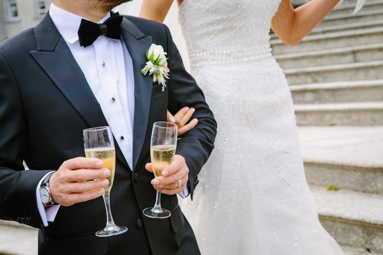 Gros plan sur un marié en smoking noir et nœud papillon, tenant deux coupes de champagne remplies, aux côtés de sa mariée dont on voit le bras et la robe en dentelle, devant des escaliers en pierre.