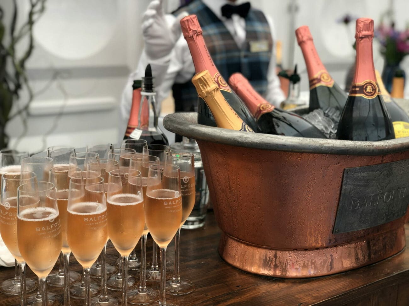 Alignement de flûtes de vin rosé effervescent à côté d'une grande vasque en cuivre remplie de glace et de bouteilles de champagne Balfour, lors d'une réception élégante.