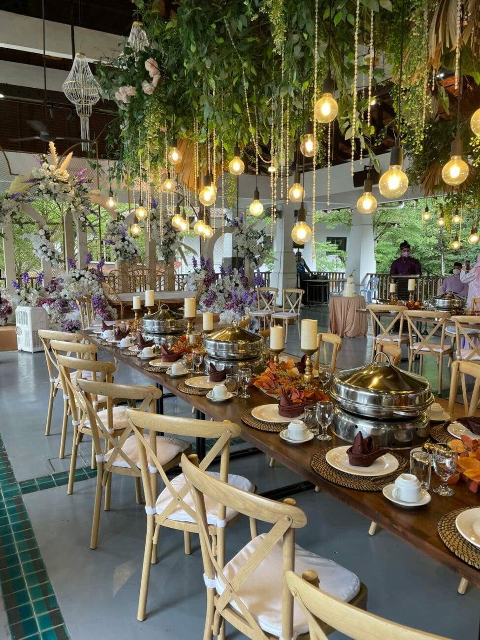 Grande salle de réception décorée de plantes tombantes et d'ampoules suspendues pour un buffet de brunch de lendemain de mariage, suggérant une sélection de vins de vins-mariages.net.