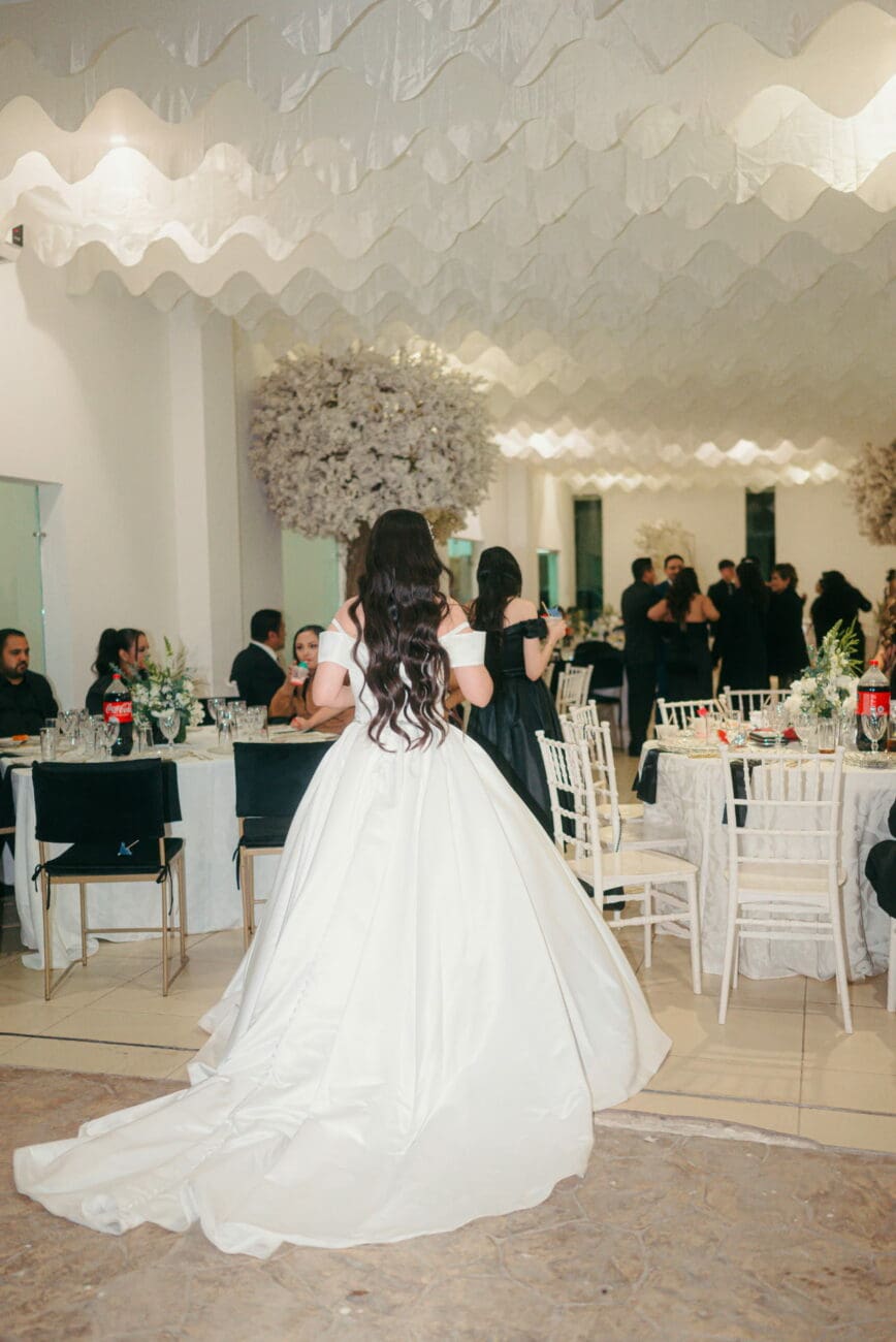 Mariée de dos en robe de bal blanche entrant dans la salle de réception du dîner, avec tables dressées et invités, par vins-mariages.net