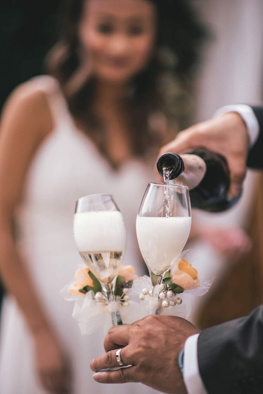 Un serveur verse du champagne dans deux flûtes décorées de fleurs et de perles, tenues par un marié, avec la mariée souriante en arrière-plan flou.