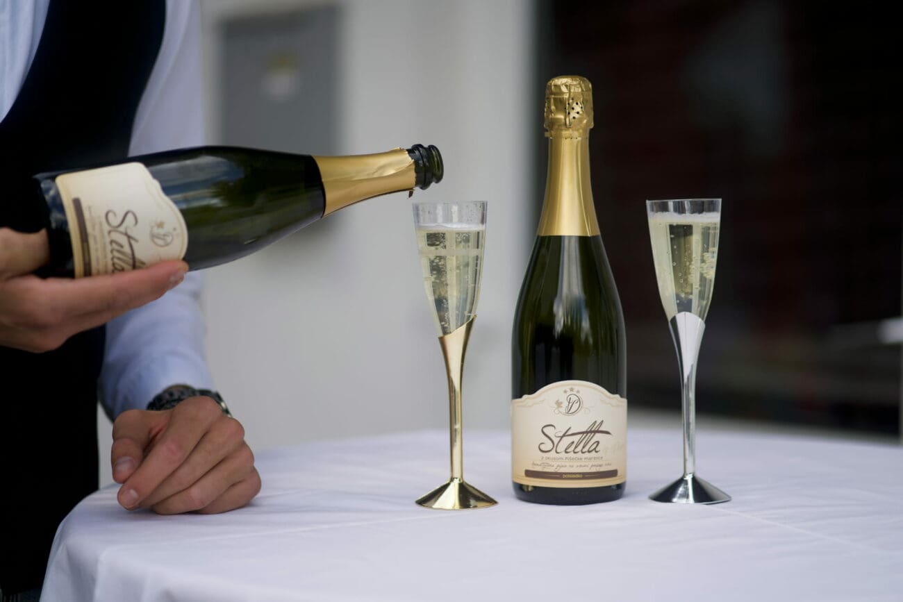 Un serveur verse du vin pétillant d'une bouteille dans une flûte dorée sur une table nappée de blanc, à côté d'une bouteille fermée et d'une flûte argentée.