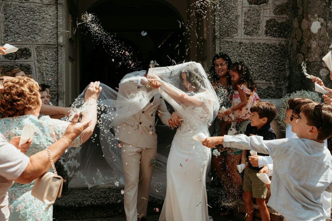 Sortie de cérémonie de mariage sous une pluie de pétales et de riz avec les mariés entourés de leurs invités devant une église en pierre.