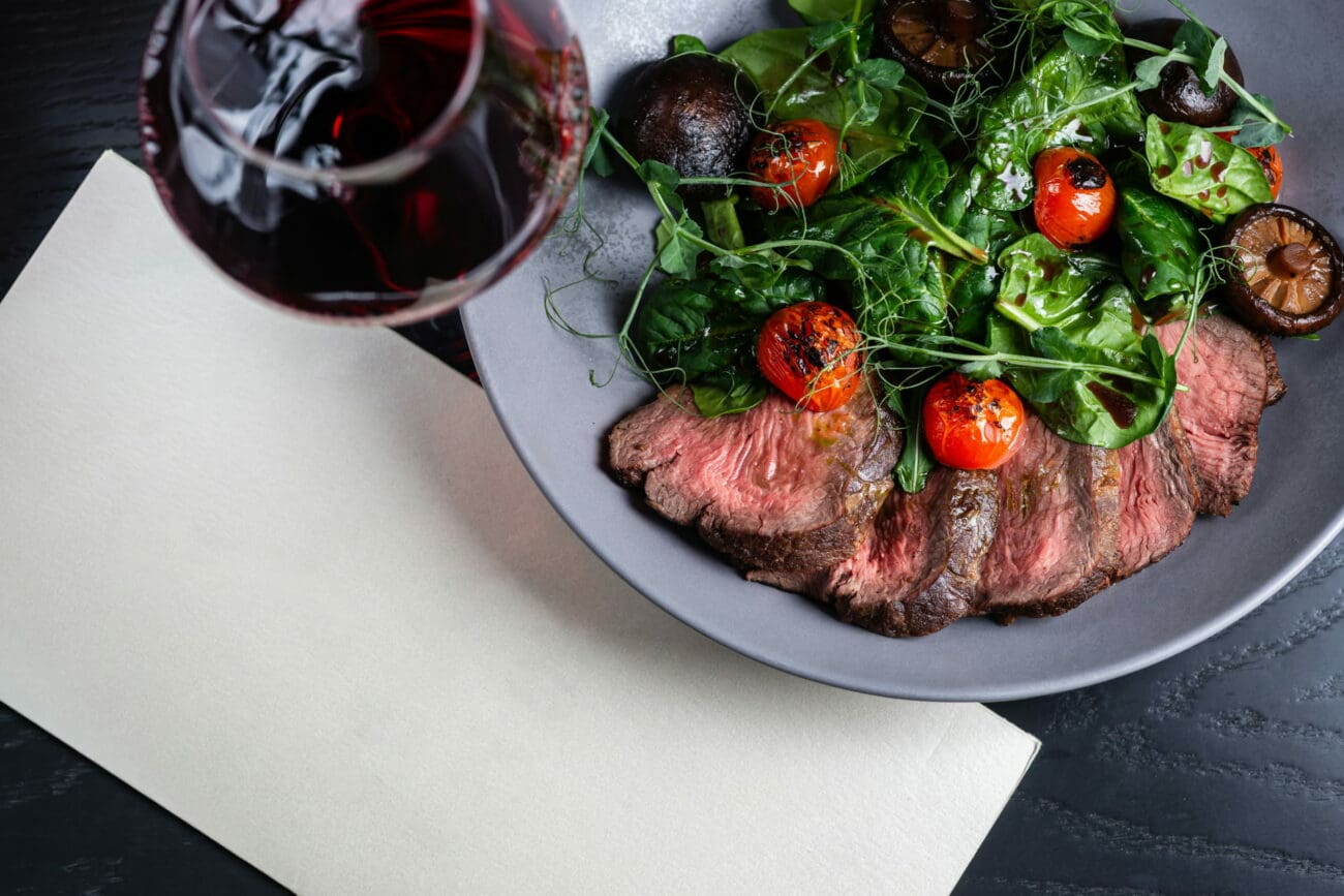 Assiette gastronomique de bœuf tranché, tomates cerises grillées et champignons, accompagnée d'un verre de vin rouge et d'un menu de mariage blanc.