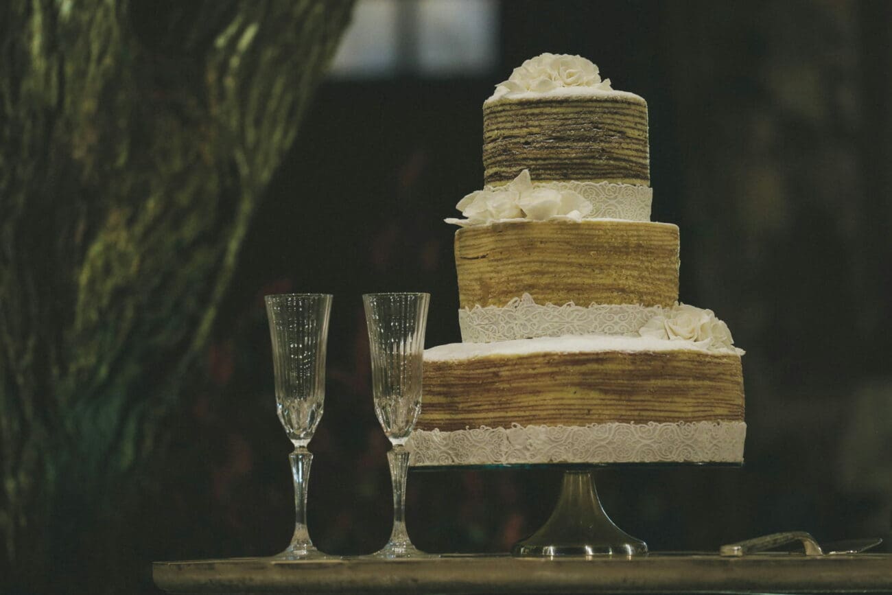 Pièce montée de mariage à trois étages décorée de dentelle blanche et fleurs, posée à côté de deux flûtes à champagne élégantes.