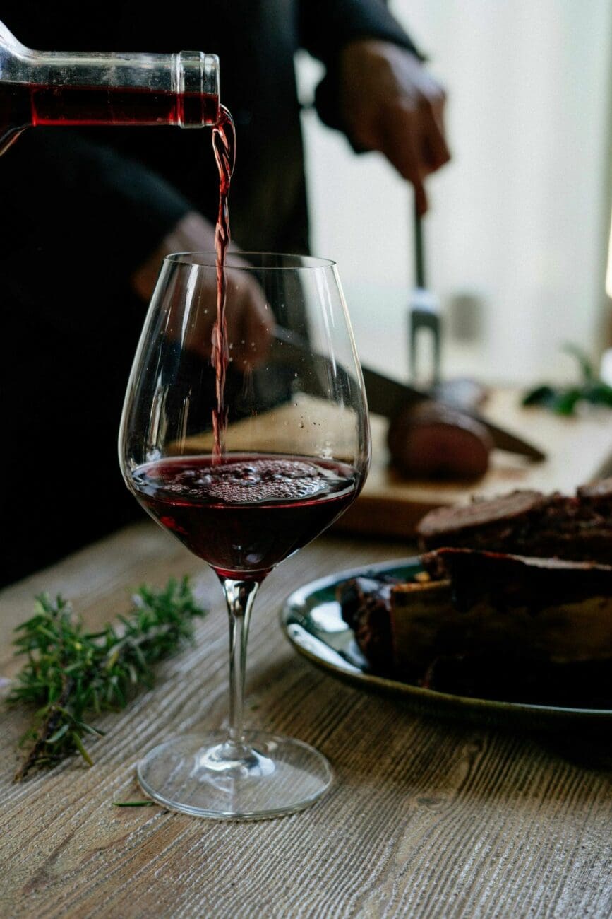 Verre de vin rouge en train d'être versé, posé sur une table en bois à côté d'une assiette de viande rôtie et de romarin, une recommandation vins-mariages.net.